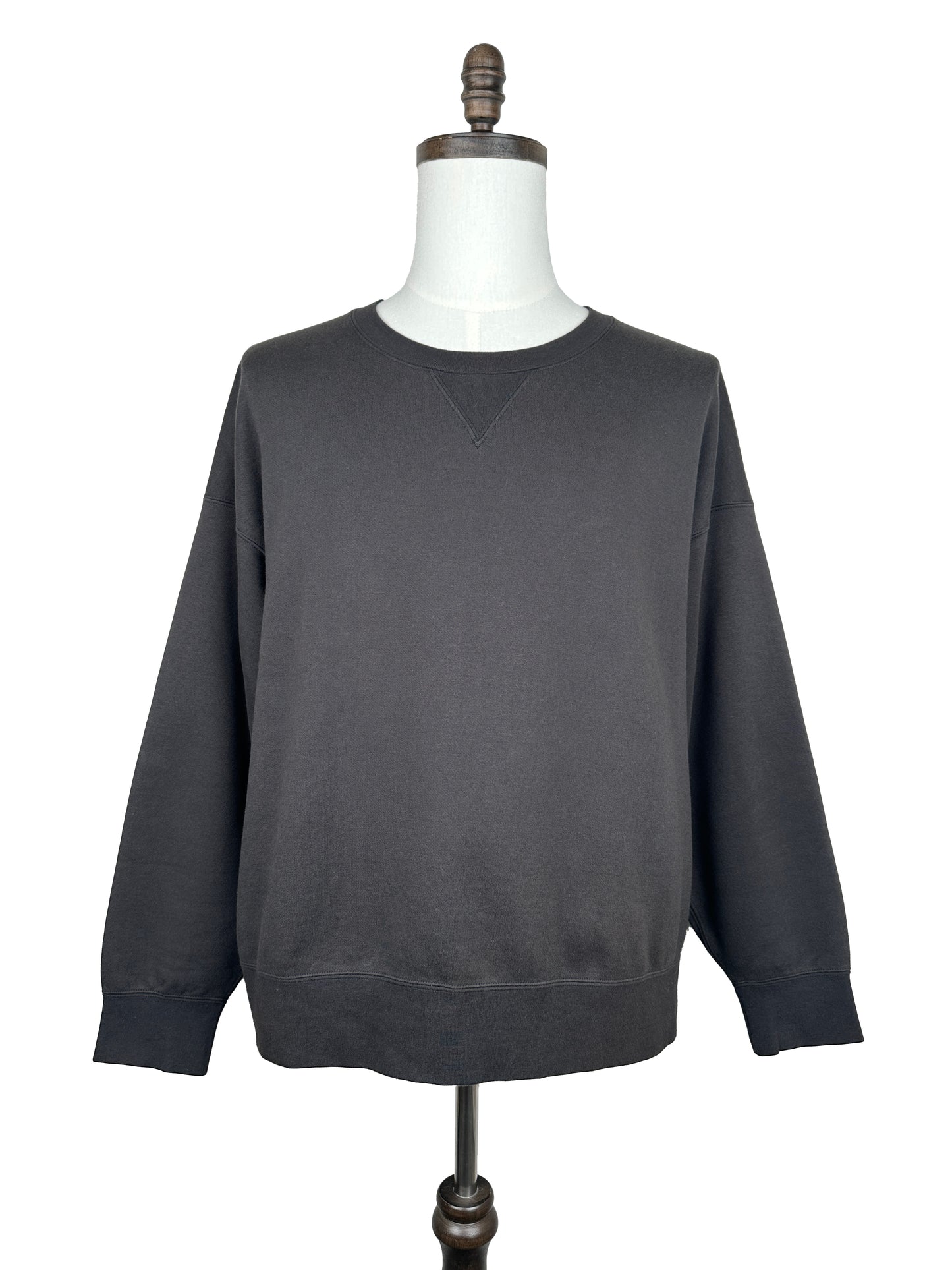 The Visvim FW23 Ultimate Jumbo SB Sweat L/S, an oversized, long-sleeved dark gray crew neck sweatshirt, is showcased on a mannequin with a white neck and wooden stand against a white background.