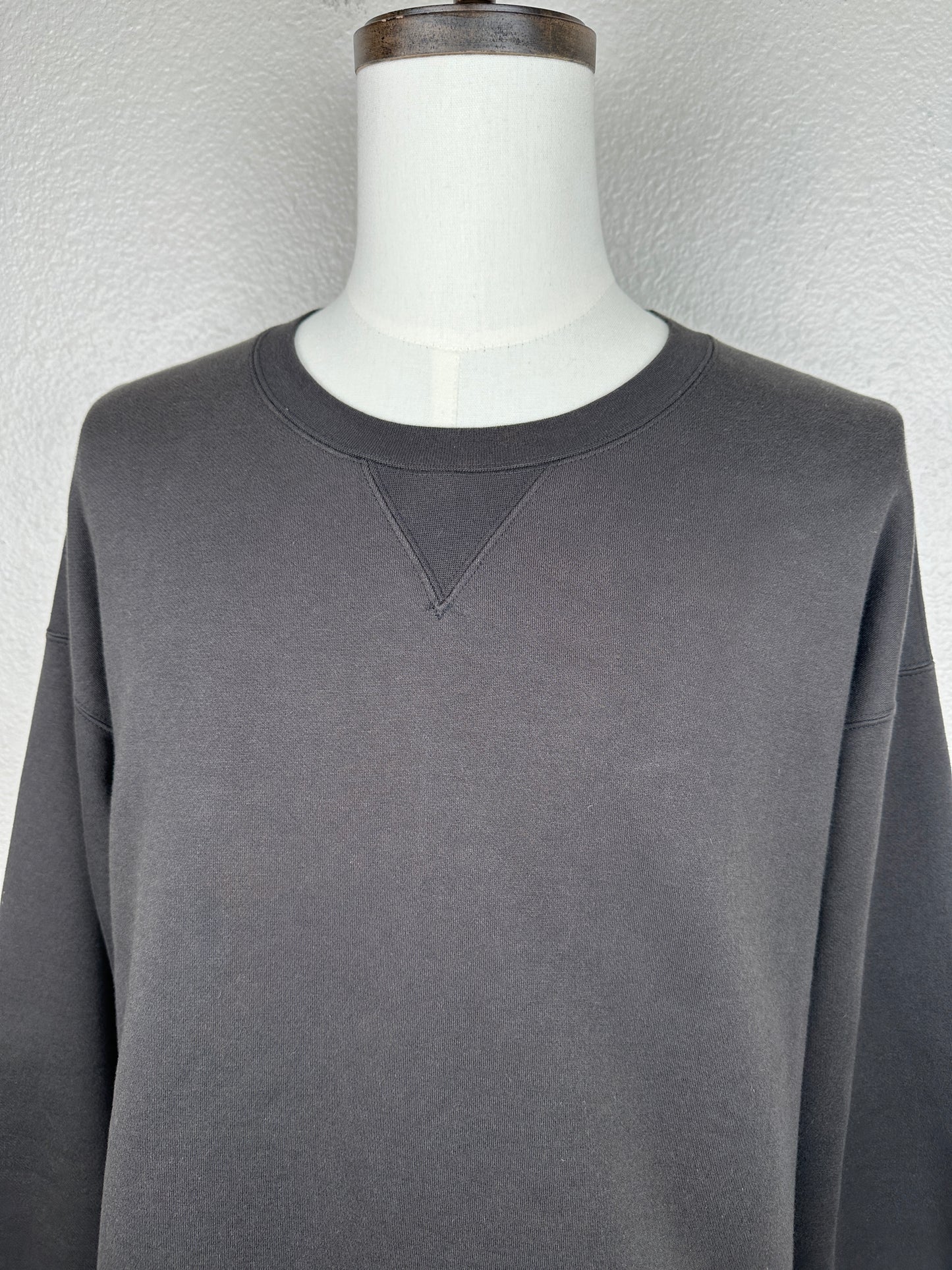 A close-up of a mannequin wearing the Visvim FW23 Ultimate Jumbo SB Sweat L/S, an oversized dark gray crewneck sweatshirt with a small stitched V detail below the neckline, set against a light textured background.