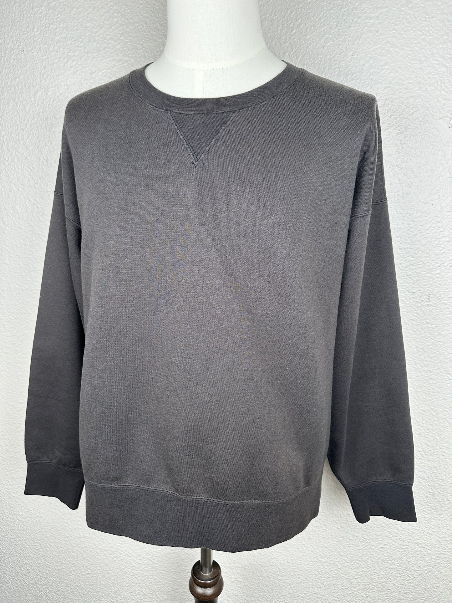 The Visvim FW23 Ultimate Jumbo SB Sweat L/S is a plain, dark gray oversized crewneck sweatshirt with long sleeves, ribbed cuffs and hem, shown on a mannequin against a light textured wall for a relaxed look.
