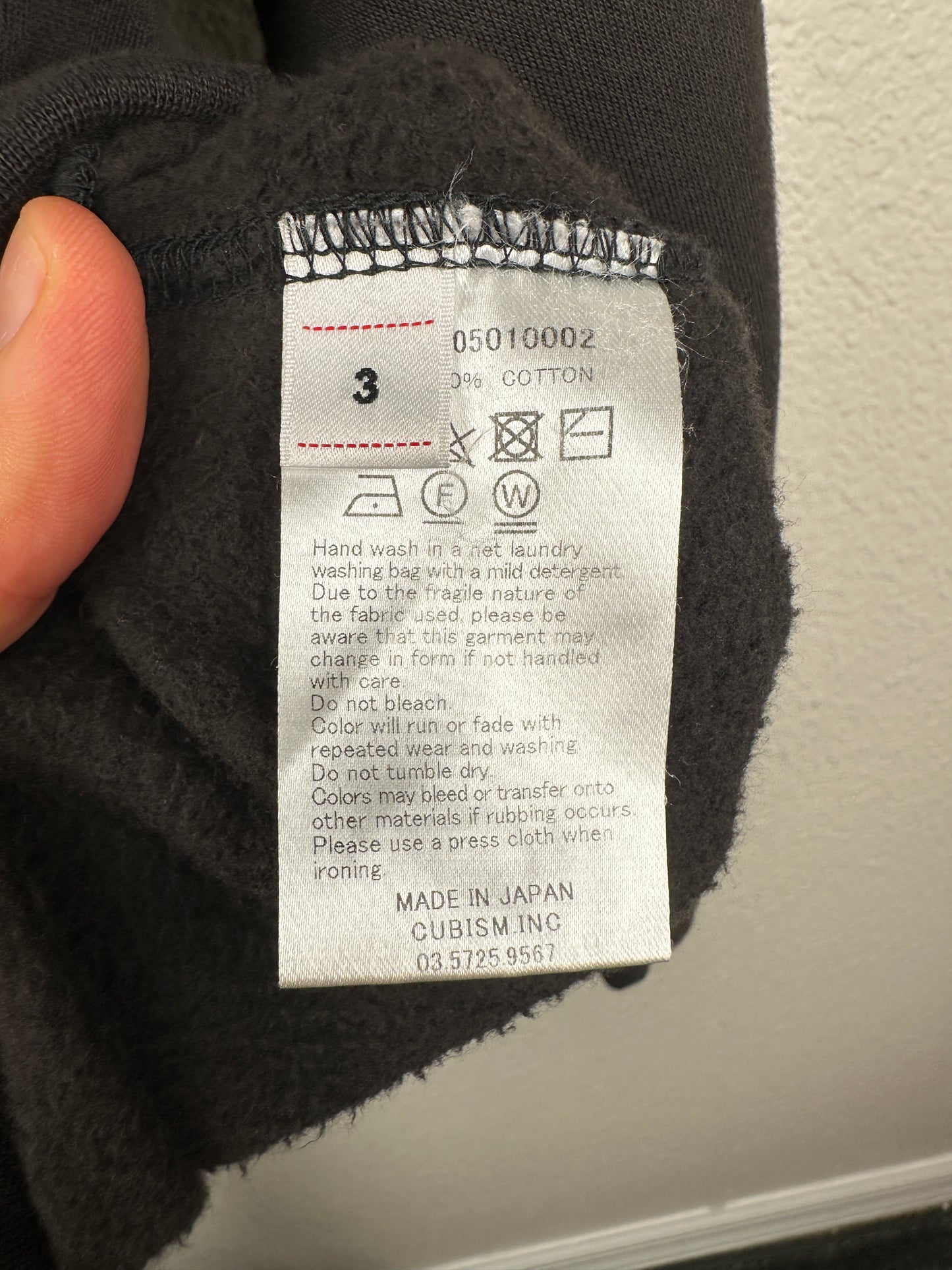 A close-up of the Visvim FW23 Ultimate Jumbo SB Sweat L/S care label on dark fabric shows washing instructions, 100% West Indian Sea Island cotton, size 3, oversized cut, Made in Japan, and CUBISM INC. contact info.