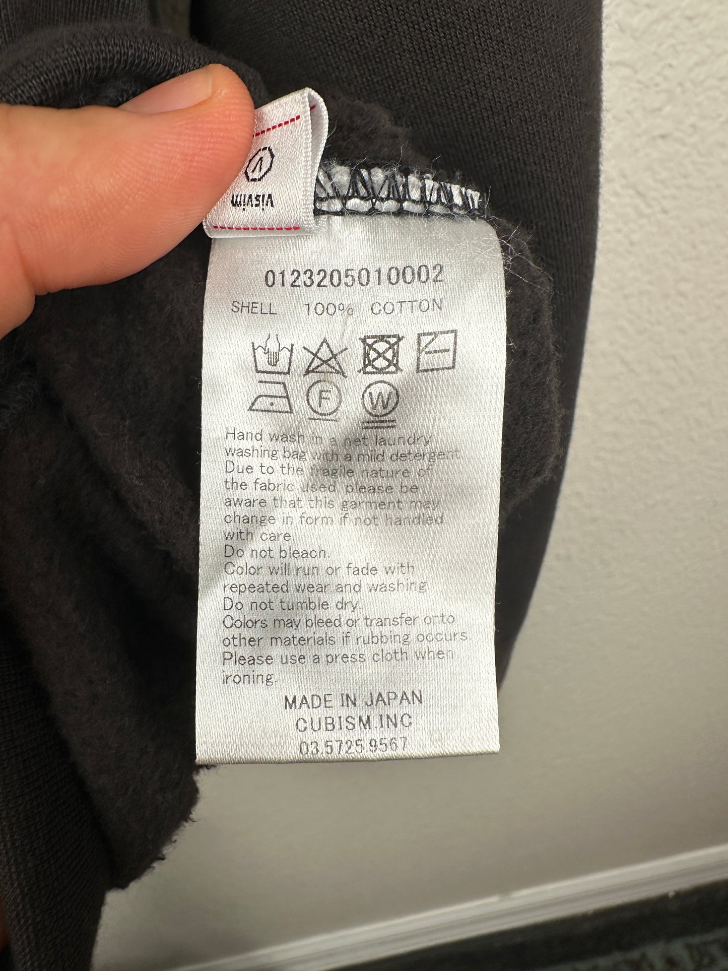 A close-up of a Visvim FW23 Ultimate Jumbo SB Sweat L/S care label shows washing symbols, instructions, and text stating it’s made with West Indian Sea Island cotton in Japan by Cubism Inc., plus a contact phone number.